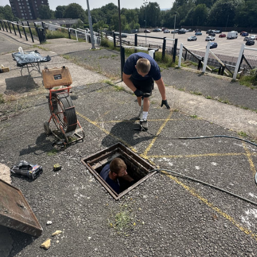 A drain unblocker down a blocked drain on a walkway with another drain engineer assisting him stood outside the drain